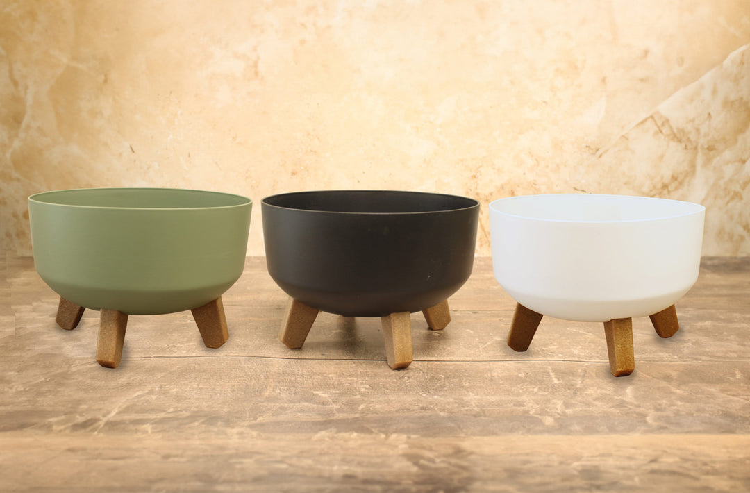 Three low plant pots with legs in green, anthracite, and white colors, displayed on a wooden surface.