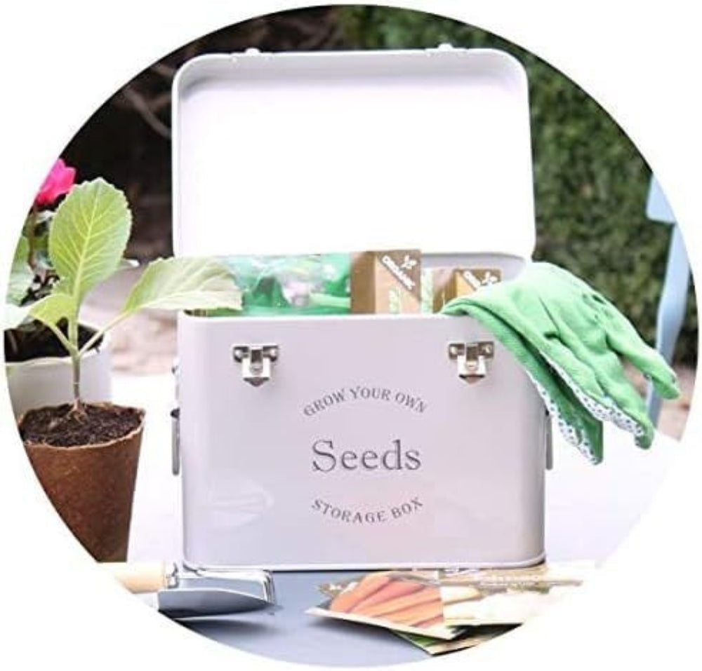 A grey seed packet storage tin with the text 'SEEDS' on the front, placed on a table with a plant pot and gardening tools in the background.