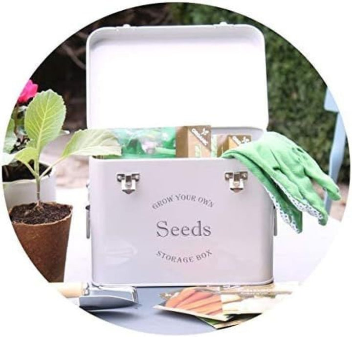 A grey seed packet storage tin with the text 'SEEDS' on the front, placed on a table with a plant pot and gardening tools in the background.