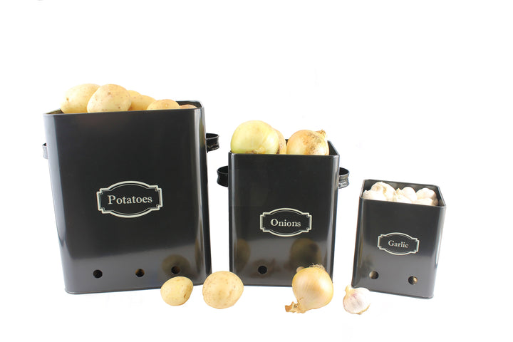 Three black kitchen storage tins labeled for potatoes, onions, and garlic, placed on a wooden countertop.
