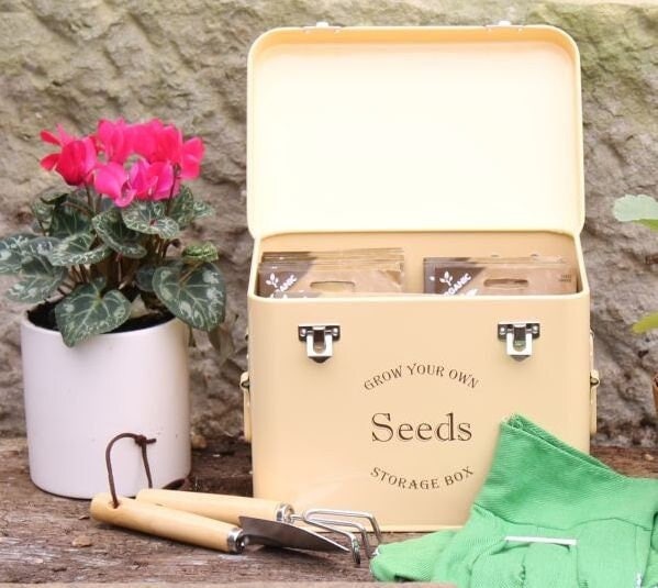 A cream-colored metal seed packet storage tin with the text 'SEEDS' on the front