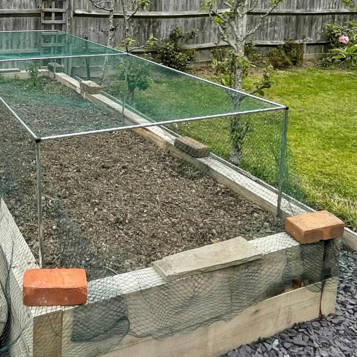A garden cage with green bird netting installed on a grassy area, containing soil and with wooden supports.