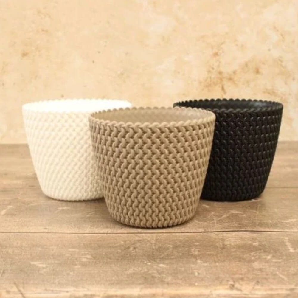Three knitted fabric flowerpots in mocha, white, and black colors, displayed on a wooden surface with a textured background.