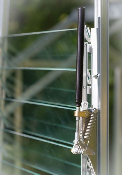 An automatic greenhouse opener attached to a greenhouse vent, with a part of a metal rod visible on the left side.
