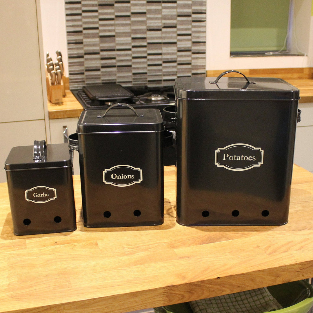Three black kitchen storage tins labeled for potatoes, onions, and garlic, placed on a wooden countertop.